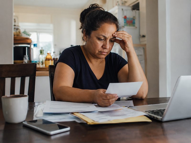 A woman at her kitchen table reviewing paperwork and bills with a laptop open.